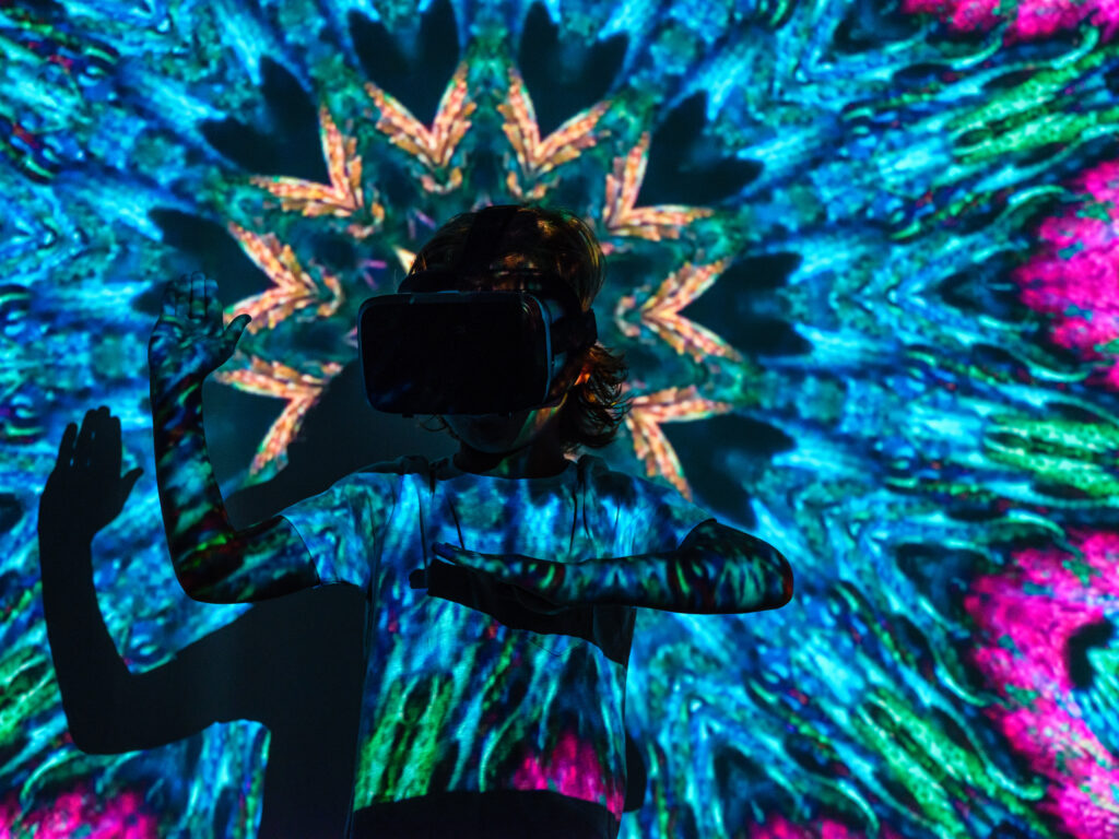 Anonymous child in VR headset playing in virtual reality while standing with raised hands under bright neon abstract colorful projector illumination