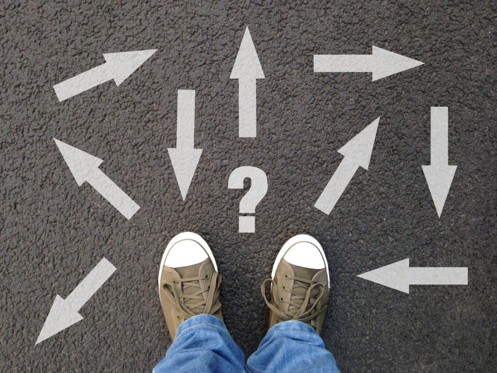 feet standing on asphalt with multitude of arrows in different directions and question mark, confusion choice chaos uncertainty concept